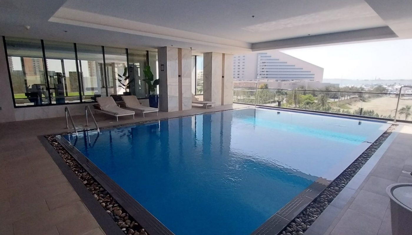 Indoor swimming pool with clear water, surrounded by a pebble border and lounge chairs, large windows offer an outside city view.