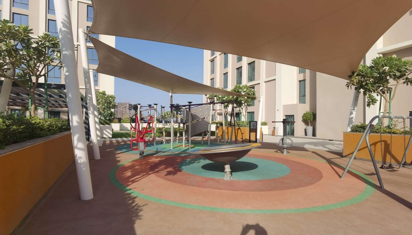 Outdoor playground with shade sails, swings, exercise equipment, and a spinning bowl, surrounded by buildings and trees on a sunny day.