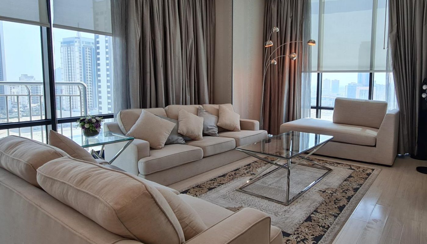 Modern living room with beige sofas, glass coffee table, floor lamp, large windows with city view, sheer curtains, and a patterned rug on wooden floor.