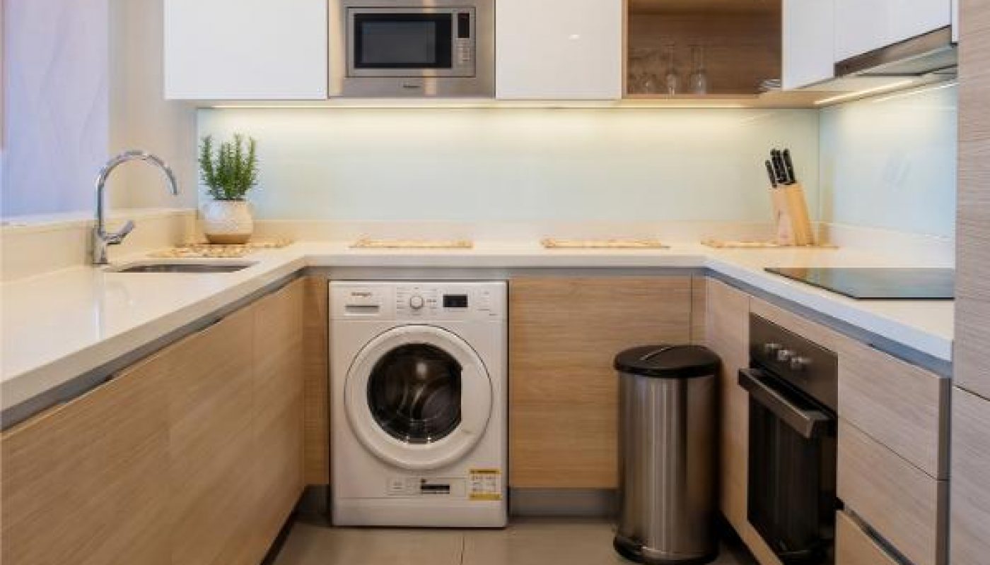 Modern kitchen with light wood and white cabinets, built-in microwave, washing machine, sink, trash bin, oven, and countertop with plant and knife holder under bright lighting.