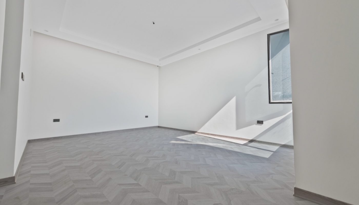 Empty, unfurnished room with white walls, a single window, and chevron-patterned gray flooring. Sunlight streams in, casting angular shadows on the floor and wall.