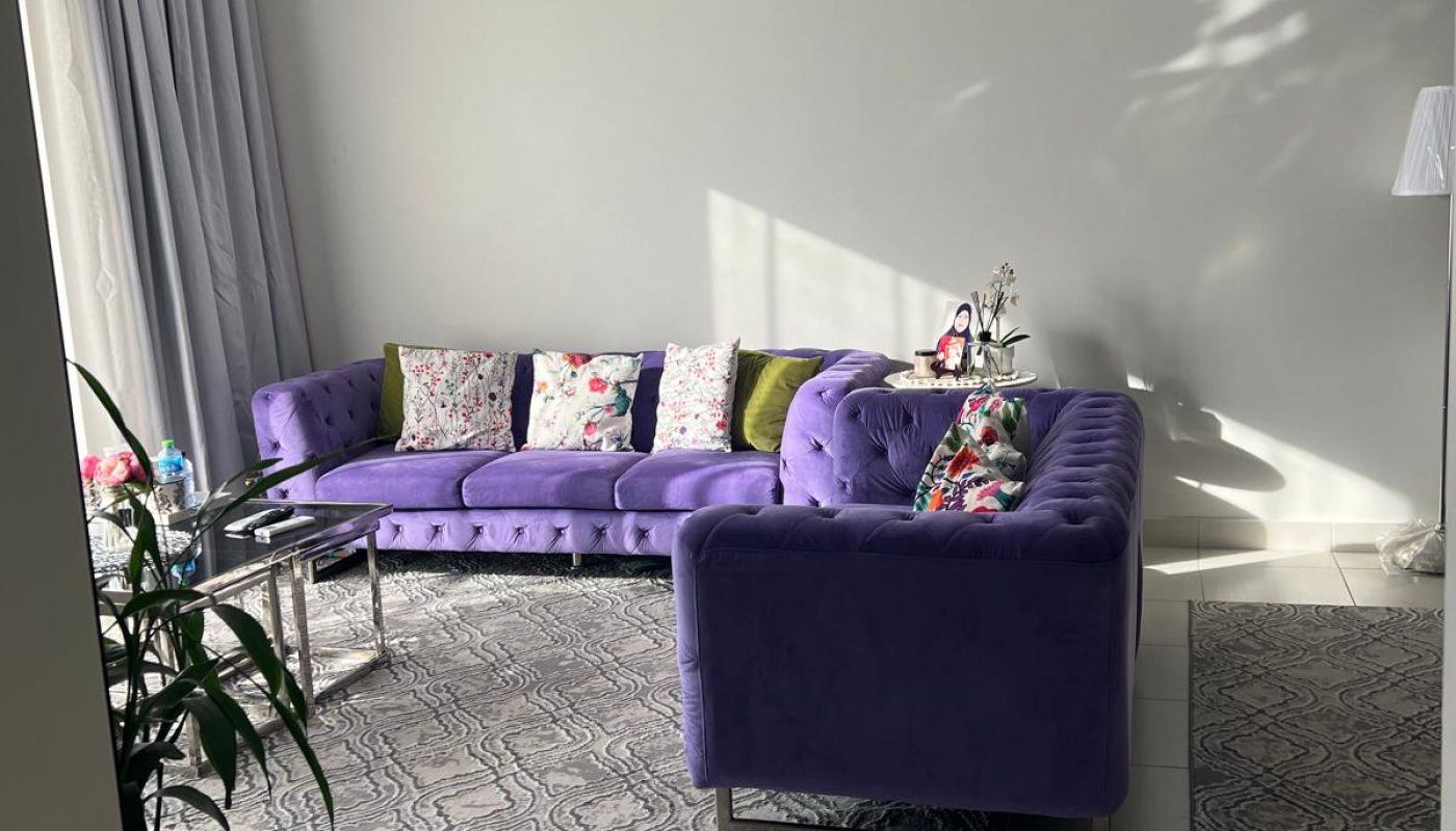 A living room with a purple sectional sofa, patterned cushions, glass coffee table, two gray rugs, potted flowers, and a wall-mounted air conditioner. Sunlight enters from a window.