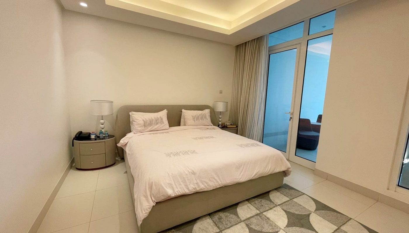 A modern bedroom with a neutral color scheme, featuring a double bed, bedside tables with lamps, an area rug, and a sliding door leading to another room. Auto Draft