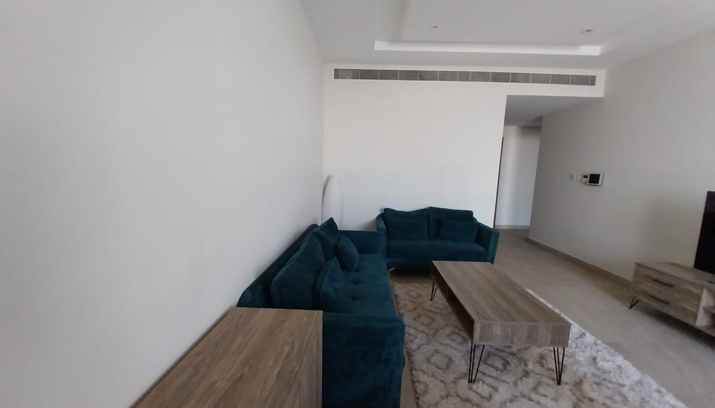 A minimalist living room with white walls, two teal sofas, a wooden coffee table, a small cabinet, and a patterned rug. An air vent is on the ceiling, and a console table is near the entrance. This flat for rent in Hidd offers modern comfort in every corner of its design.