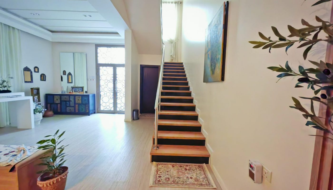 A bright hallway with a straight wooden staircase, a small rug at its base, potted plants, wall art, and a glass entry door are visible.