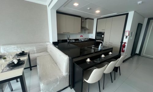 Modern kitchen in a fully furnished 2 BR apartment with white cabinets and black countertops, including an island and dining area with chairs still wrapped in protective plastic.