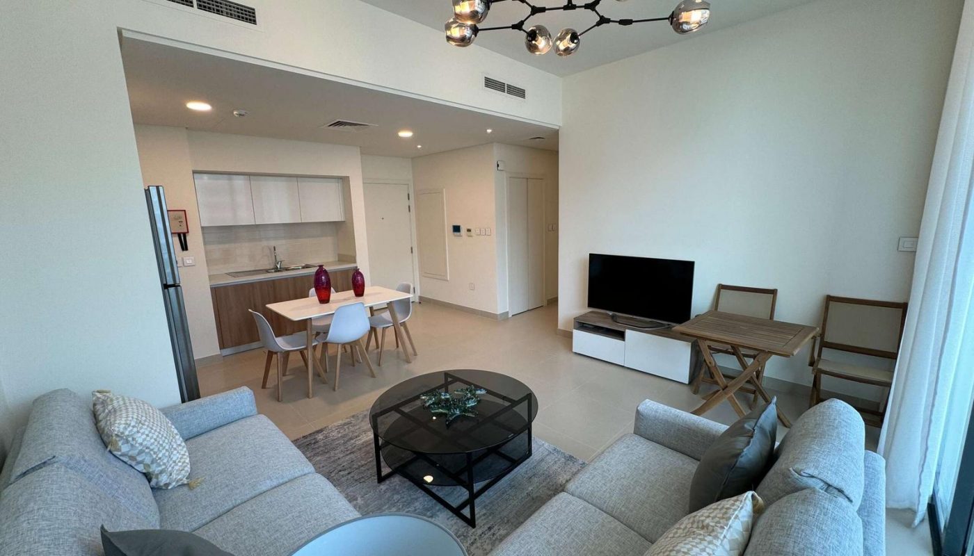 Modern living room with two gray sofas, glass coffee table, TV, dining area with four chairs, kitchenette, and contemporary light fixture. Large windows provide natural light.