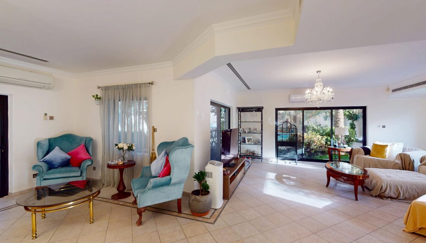 Spacious living room with blue and beige armchairs, glass coffee tables, a sofa, large windows, tile flooring, and indoor plants. Natural light fills the area.