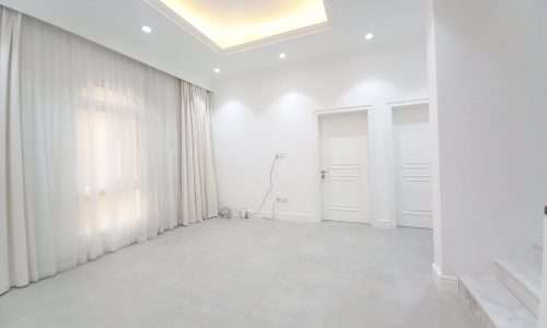 A minimalist room in a luxury villa for rent in Janabiya, featuring white walls, a recessed ceiling with lights, floor-to-ceiling curtains, two closed white doors, and some electrical wires on the floor.