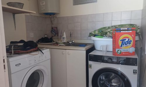 A laundry room with a washing machine, a dryer, a sink, shelves with various items, and a box of Tide detergent on the counter.