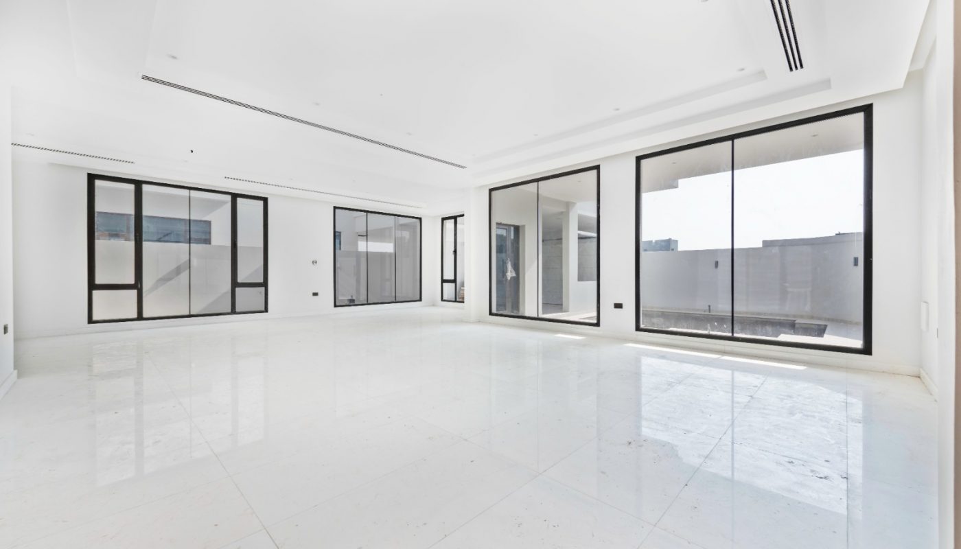 Spacious empty room with white floors and walls, large floor-to-ceiling windows, and ample natural light coming in from outside.