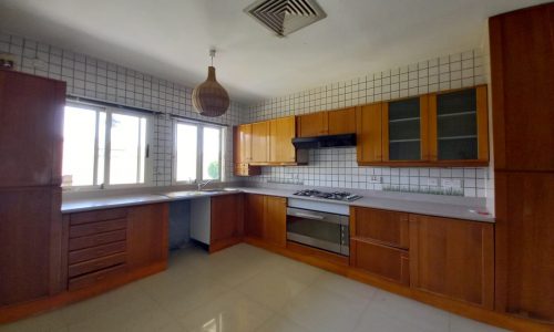 A spacious kitchen with wooden cabinets, white tiled walls, a built-in stovetop and oven, large windows, and a hanging wicker light fixture.