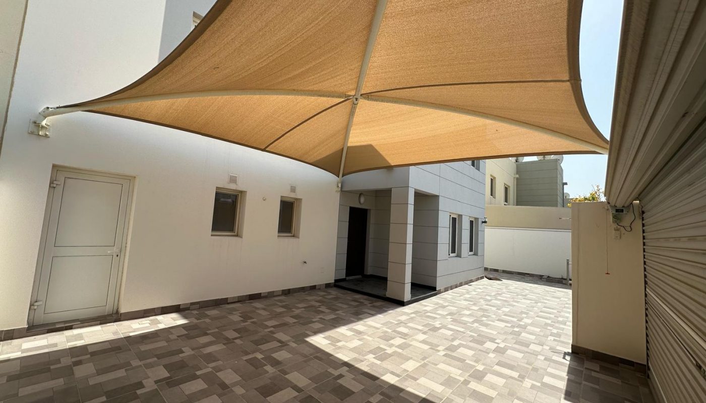 Spacious tiled courtyard with a large beige shade sail, adjacent to a modern building with white and gray walls and several windows and doors.