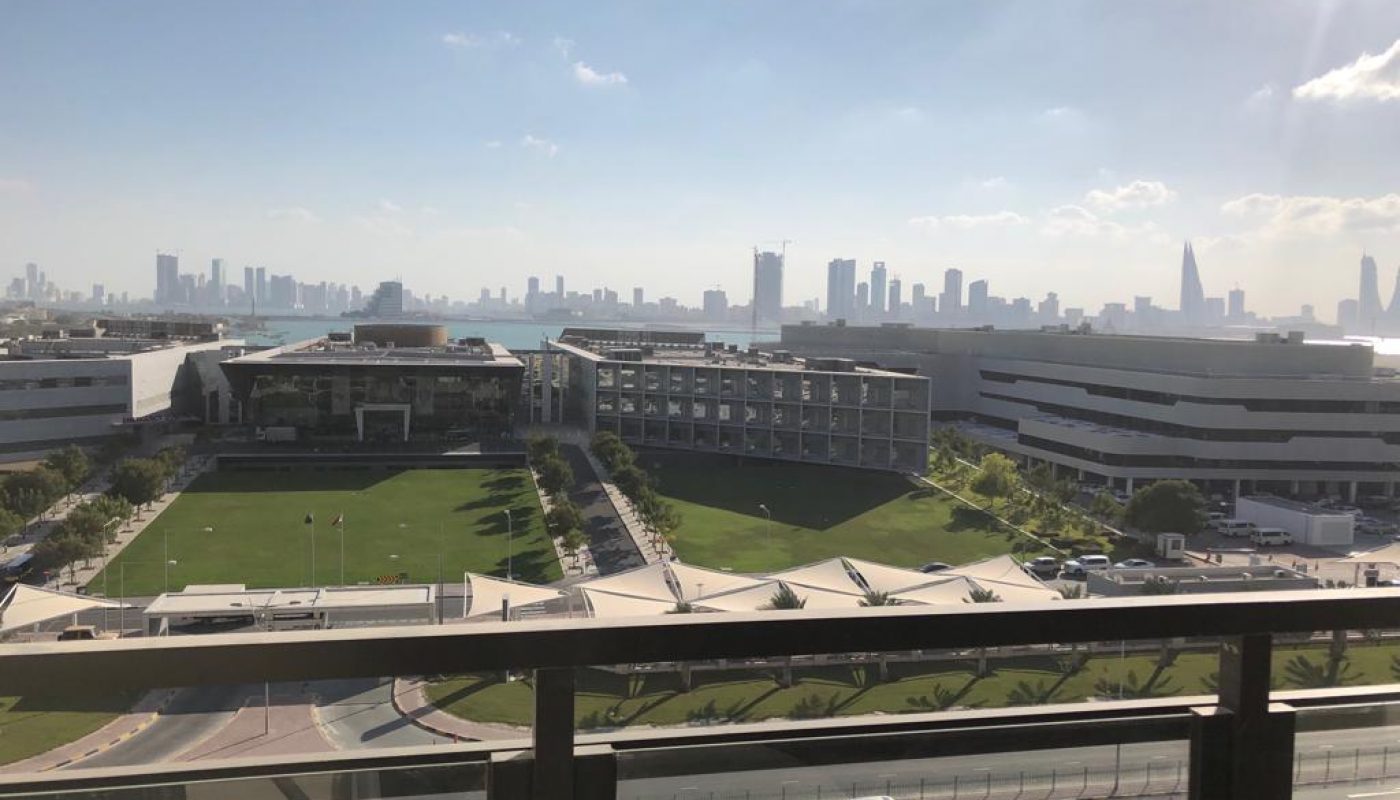 View from a balcony overlooking a modern campus with green lawns, surrounding buildings, and a city skyline under a partly cloudy sky.