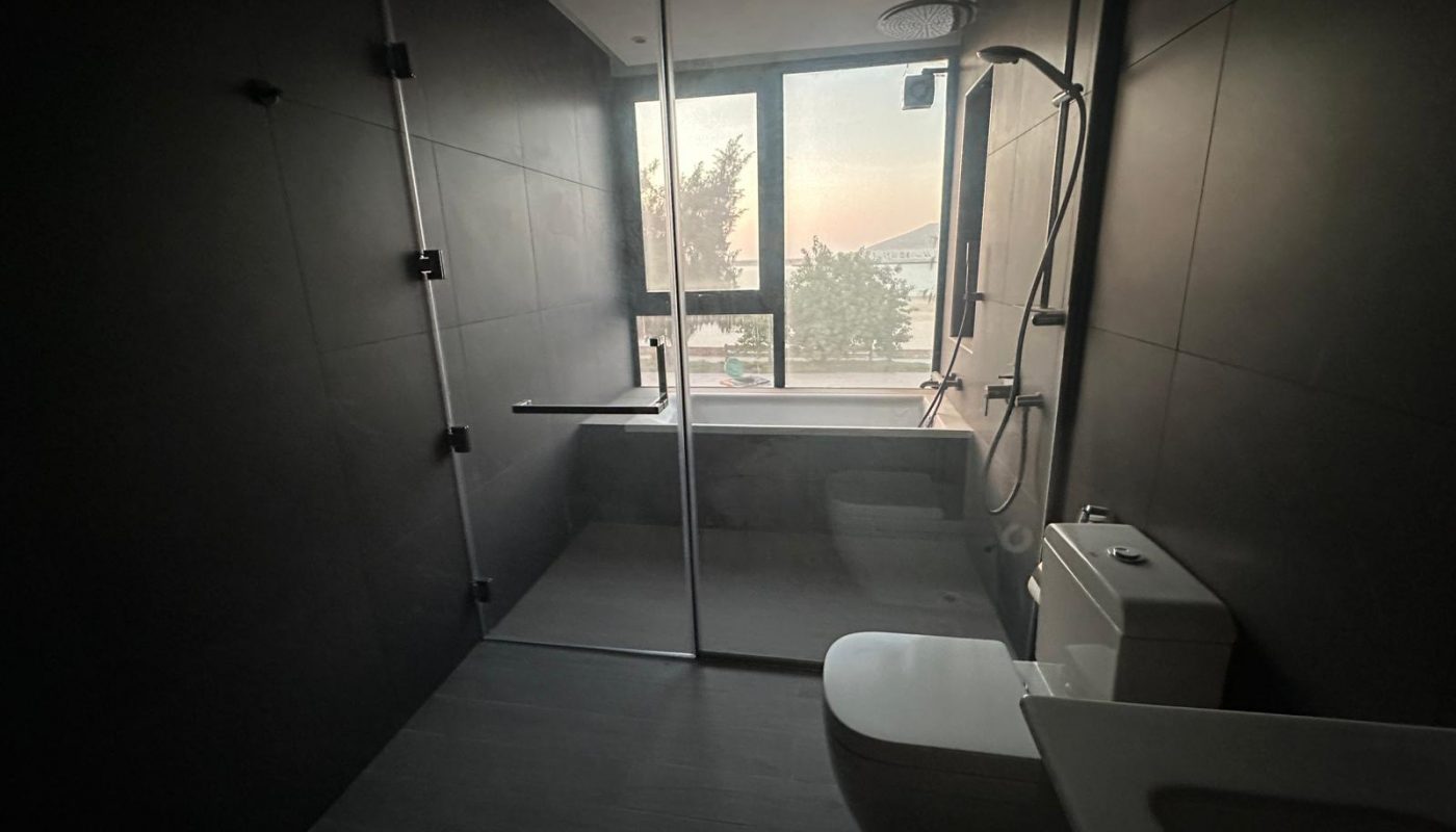 Modern bathroom with dark tiles, glass shower enclosure, white toilet, and a window letting in natural light.