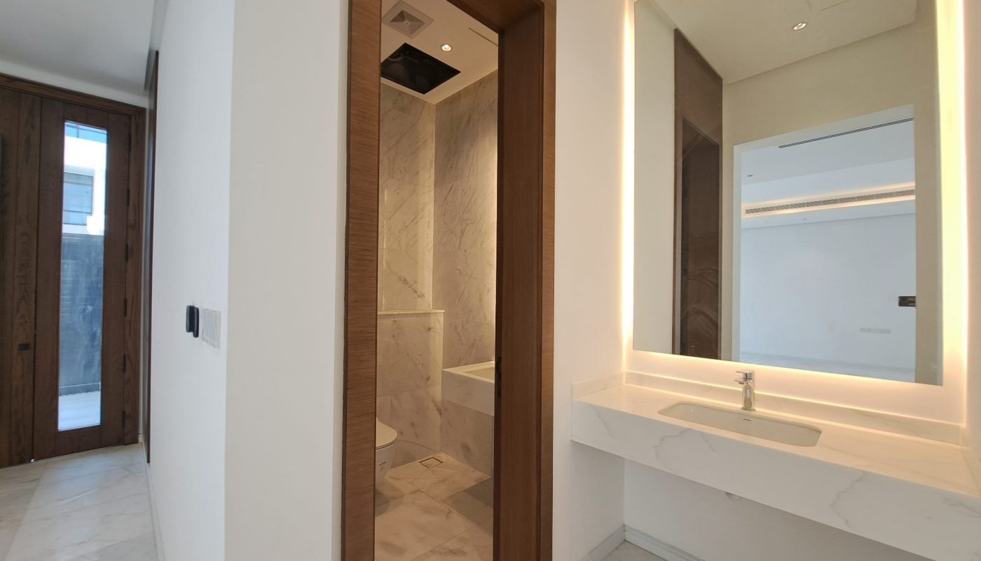 Modern bathroom with a large illuminated mirror, white countertop with sink, and a separate toilet area featuring marble walls and floors.