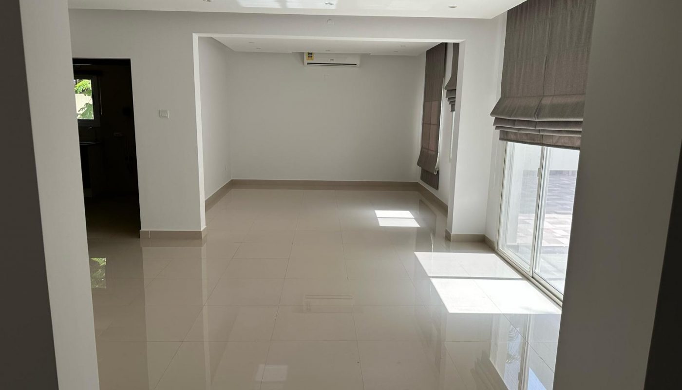 Empty room with white walls, shiny tiled floor, large windows with curtains, and a wall-mounted air conditioner. Natural light enters through the windows.