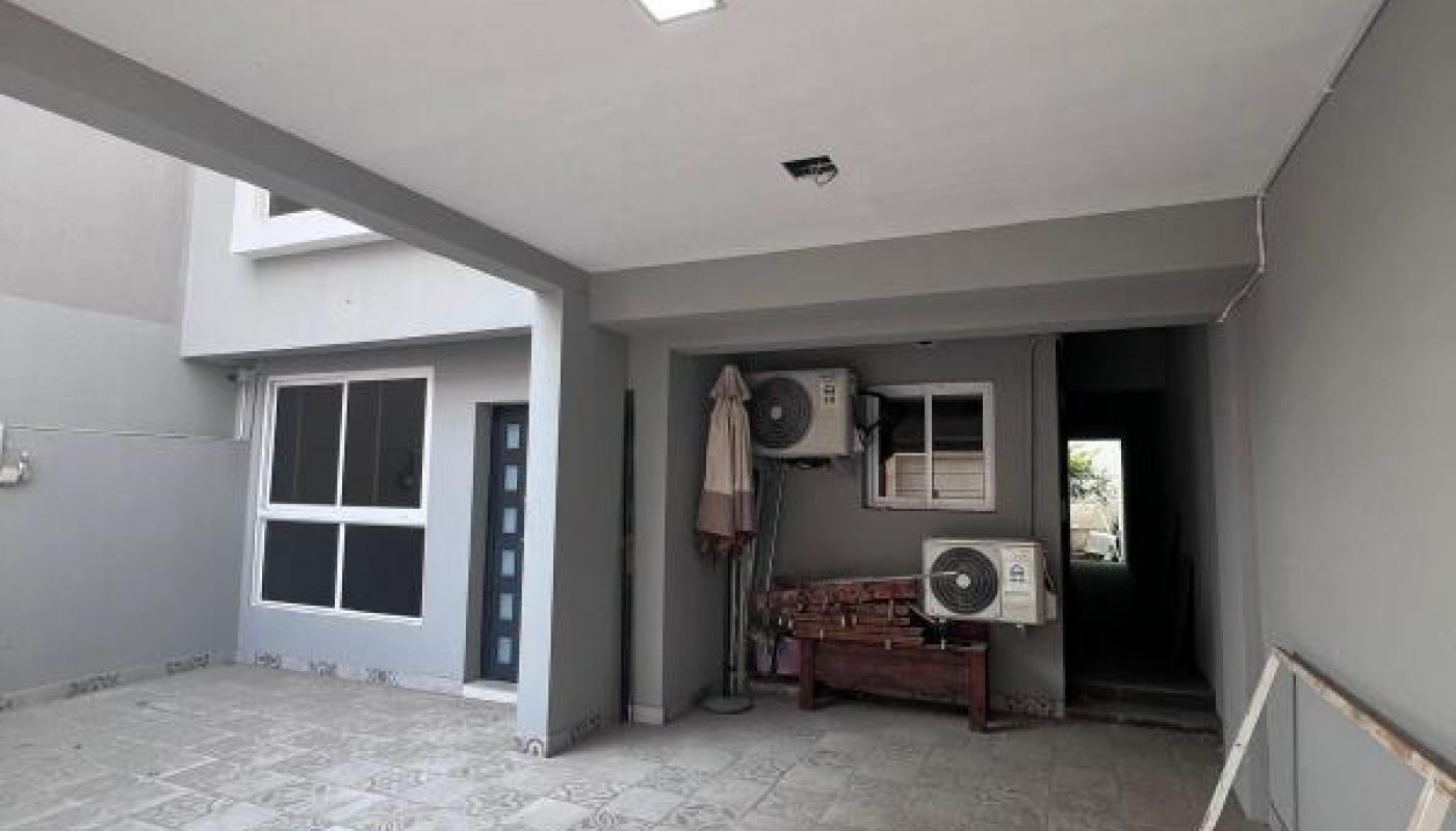 A covered outdoor area with tiled flooring, a wooden bench, two air conditioning units, a window, a door, and a coiled garden hose on the ground.