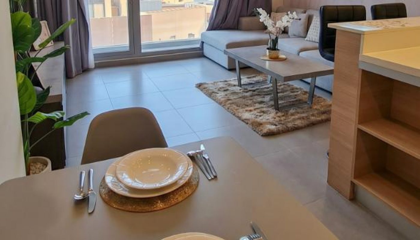 Modern apartment living room and dining area with large window, set dining table for two, sofa, coffee table, wall art, and city view outside.