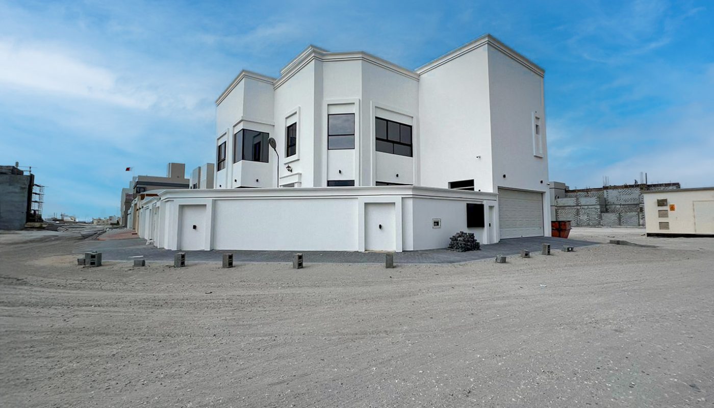A white two-story building with large windows stands on a sandy, open lot under a blue sky with scattered clouds.