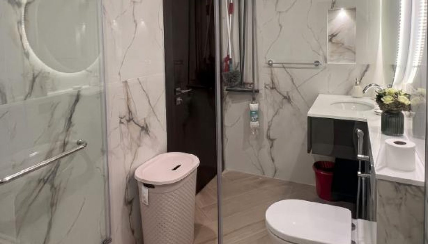 Modern bathroom with marble-patterned walls, glass shower enclosure, round mirror, wall-mounted toilet, laundry basket, small shelf with plant, and cleaning tools behind the door.