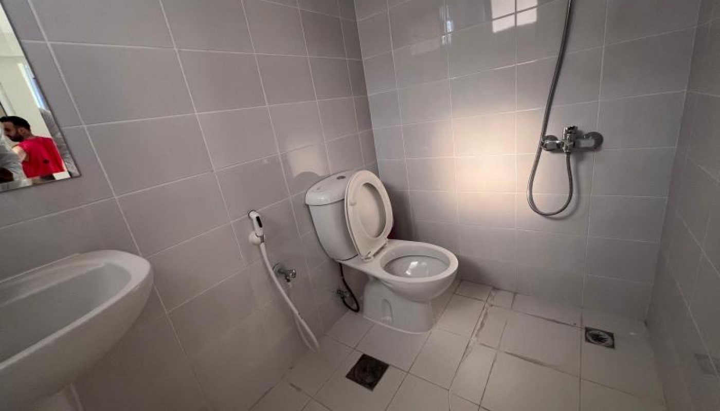A bathroom with a toilet, handheld bidet, sink, and wall-mounted showerhead, all set against light gray tiled walls and floor.