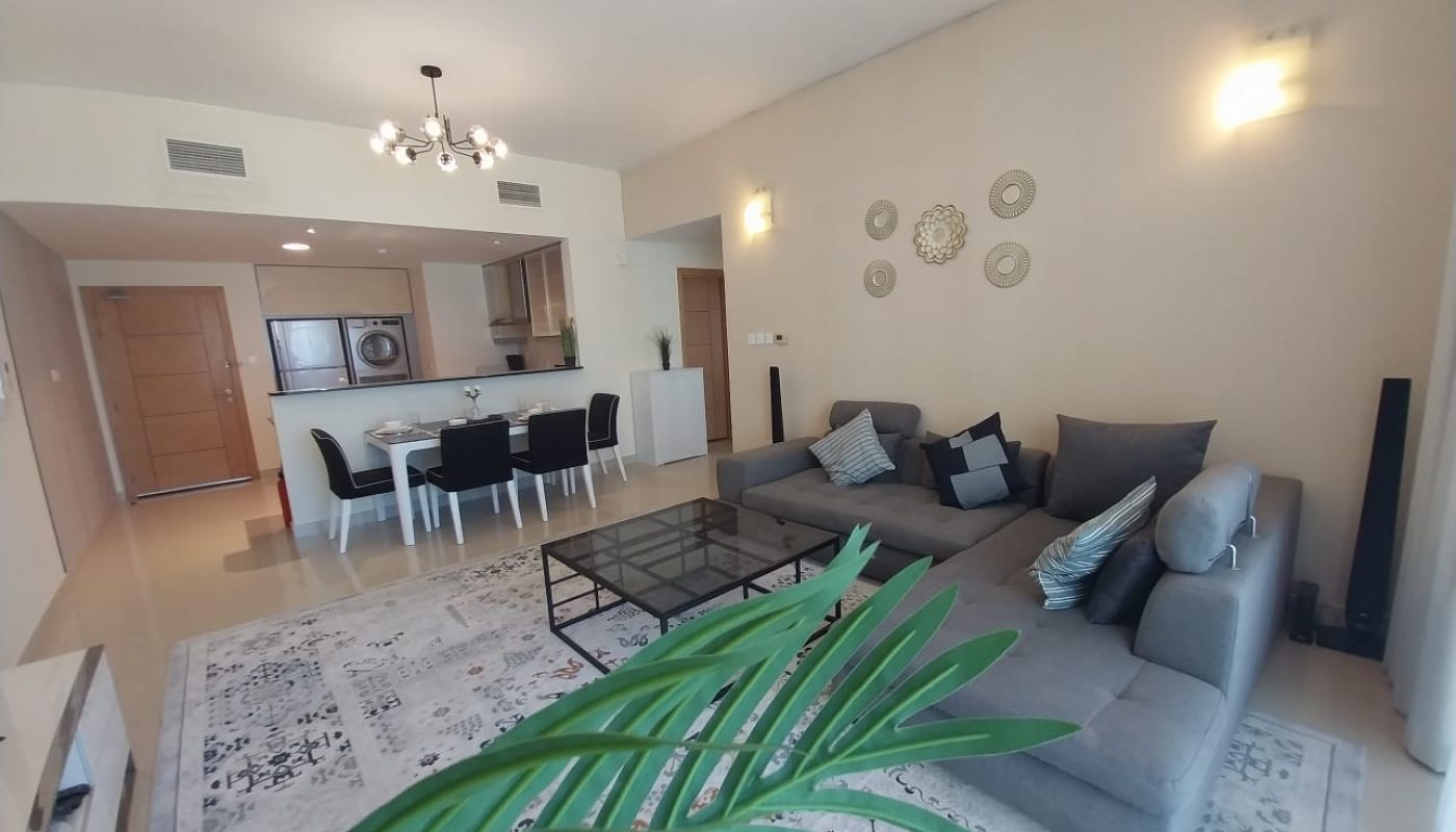 Modern living room with a gray sectional sofa, glass coffee table, dining area, open kitchen, decorative wall plates, and a potted plant in the foreground.