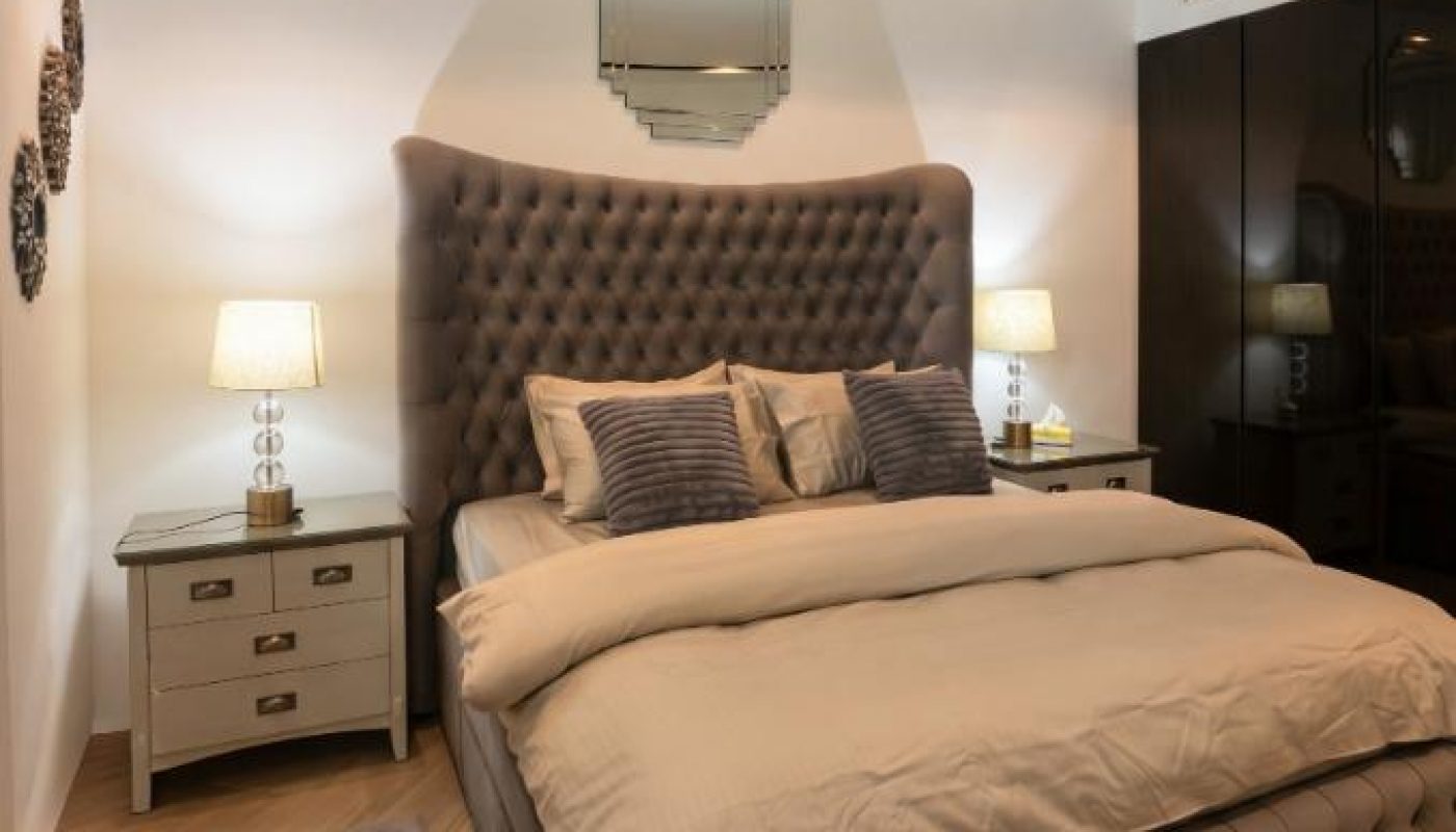 A modern bedroom with a tufted bed, beige bedding, two bedside tables with lamps, a mirror above the headboard, and dark wardrobes on the right.