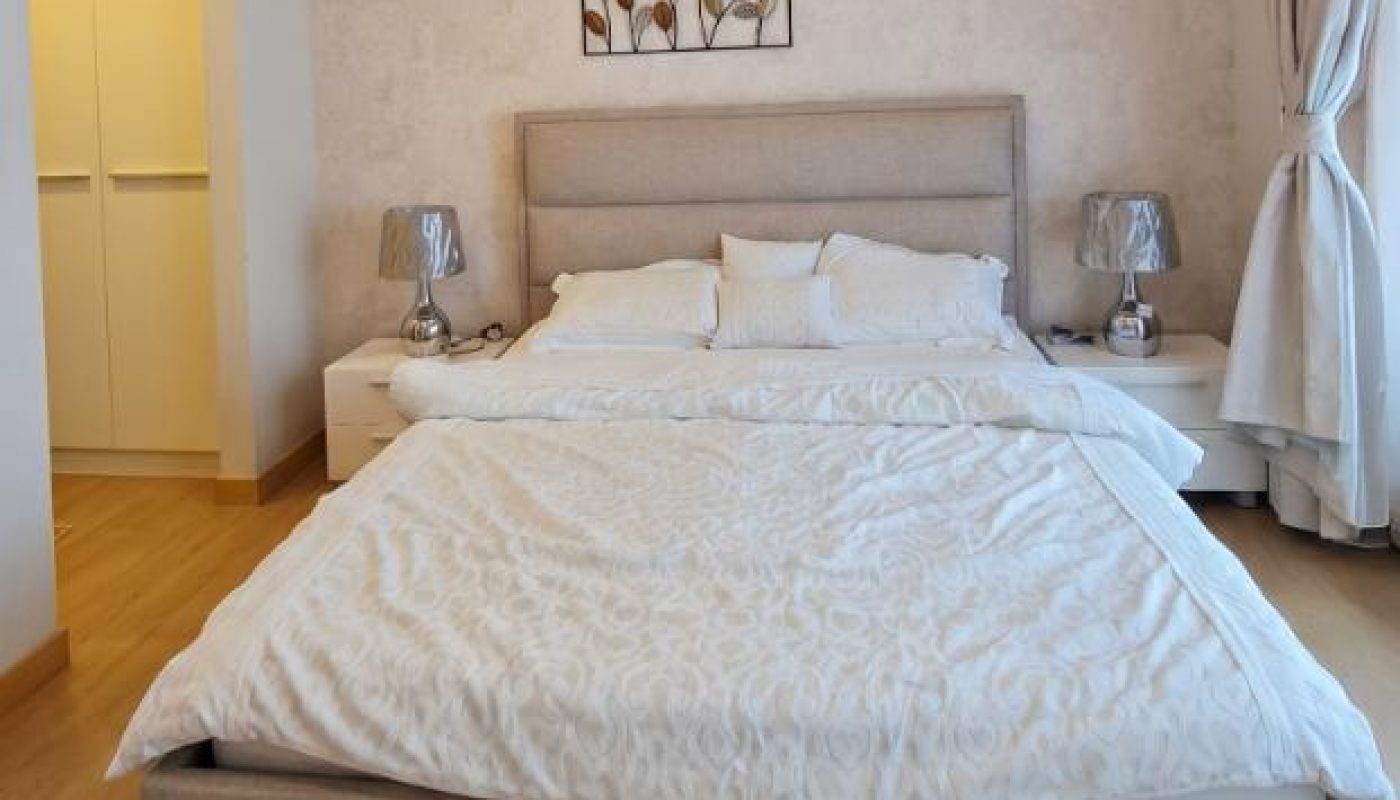 Modern bedroom with a beige upholstered bed, white bedding, two bedside tables with lamps, wall art above the headboard, and a window with curtains.