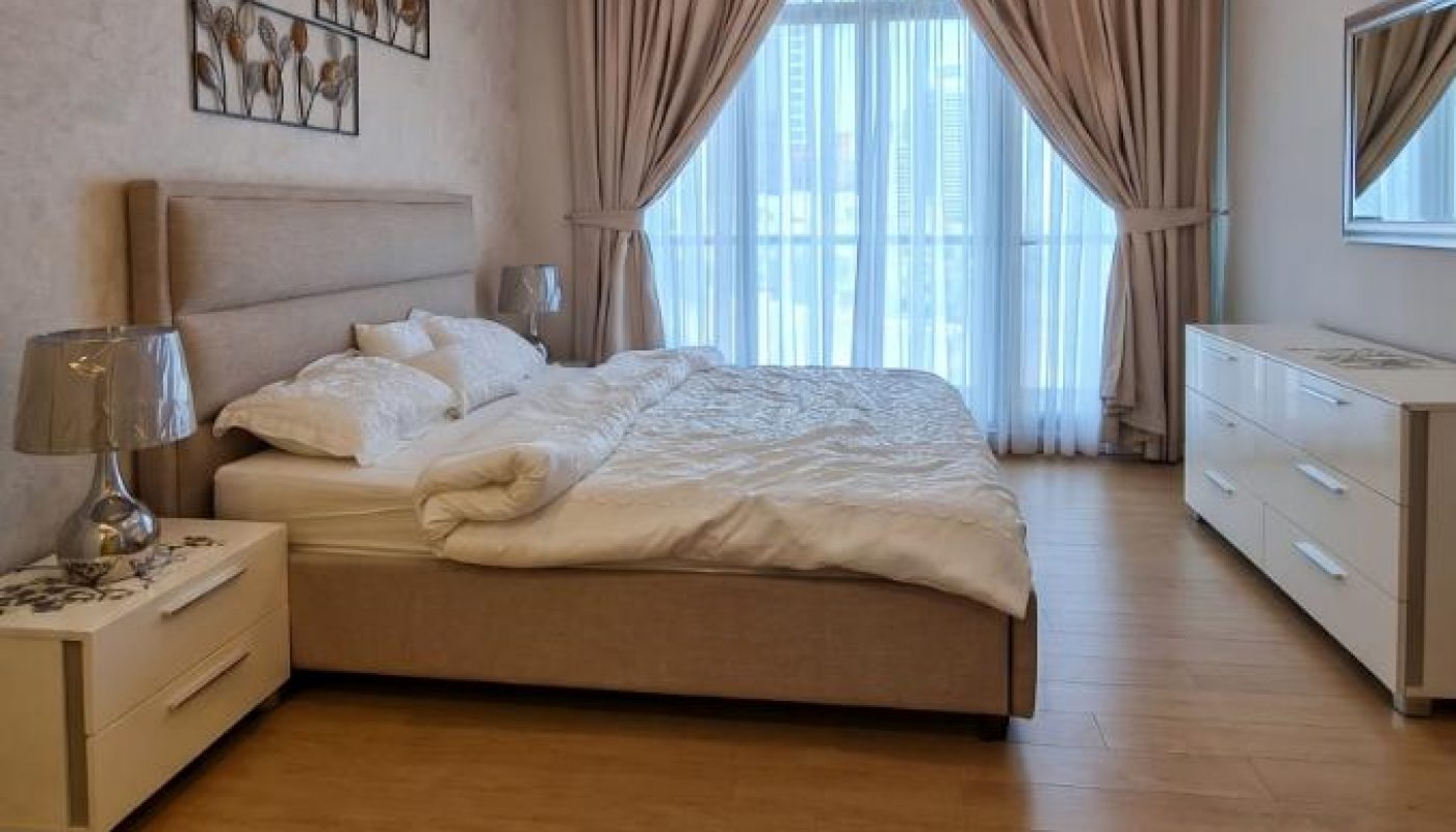 Modern bedroom with a large bed, two bedside tables, a dresser with a mirror, wooden floor, and large window with beige curtains letting in natural light.