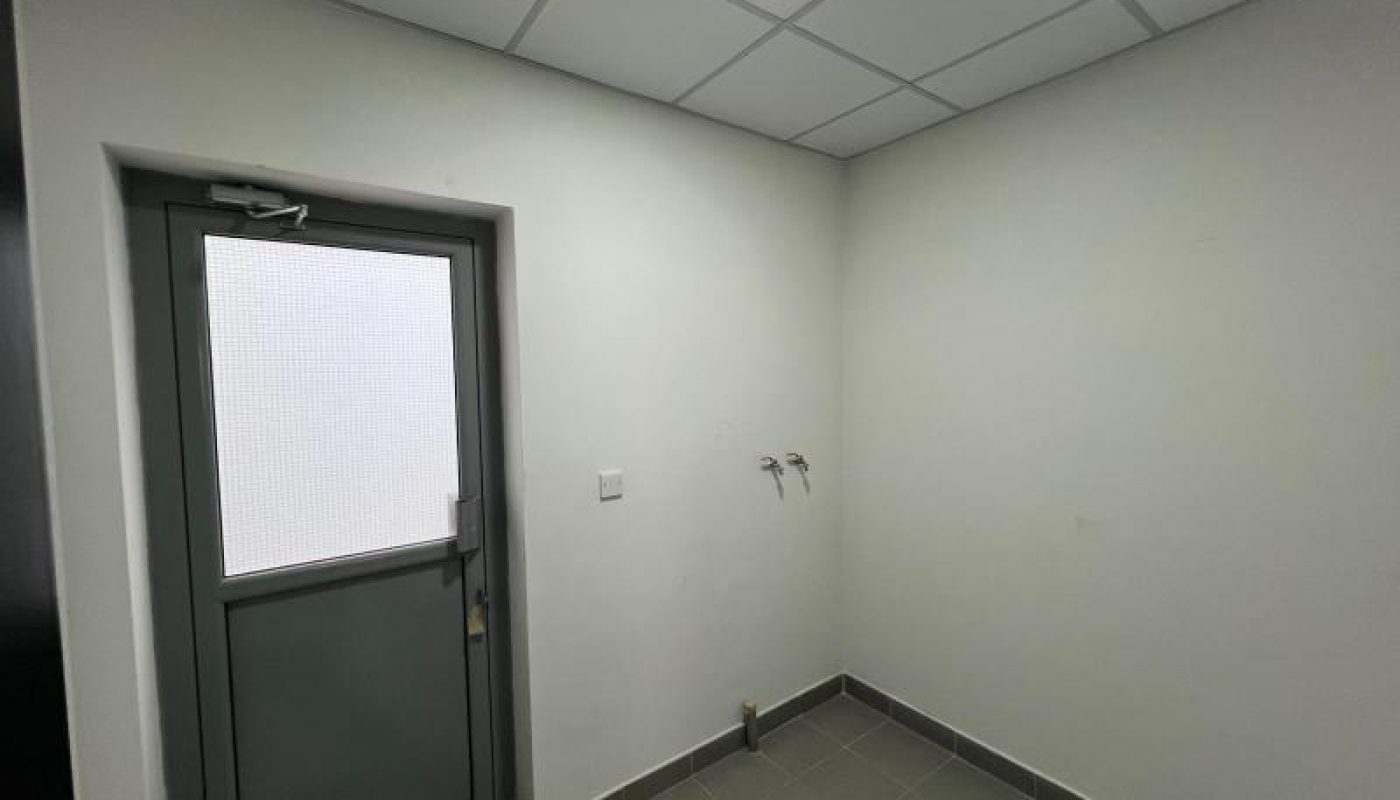 Empty, small room with tiled floor, white walls, a closed gray door with a frosted window, ceiling light, and two wall-mounted water outlets.