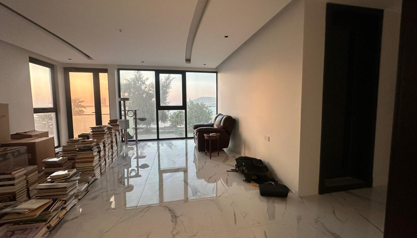 A spacious, modern room with marble floors, large windows, stacks of books, a leather armchair with a footrest, and a few bags on the floor near the entryway.