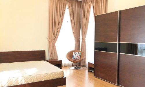 A bedroom with a wooden bed frame and matching nightstand, large windows with beige curtains, a cushioned chair, and a dark wood wardrobe. The room has light-colored walls and wooden flooring.