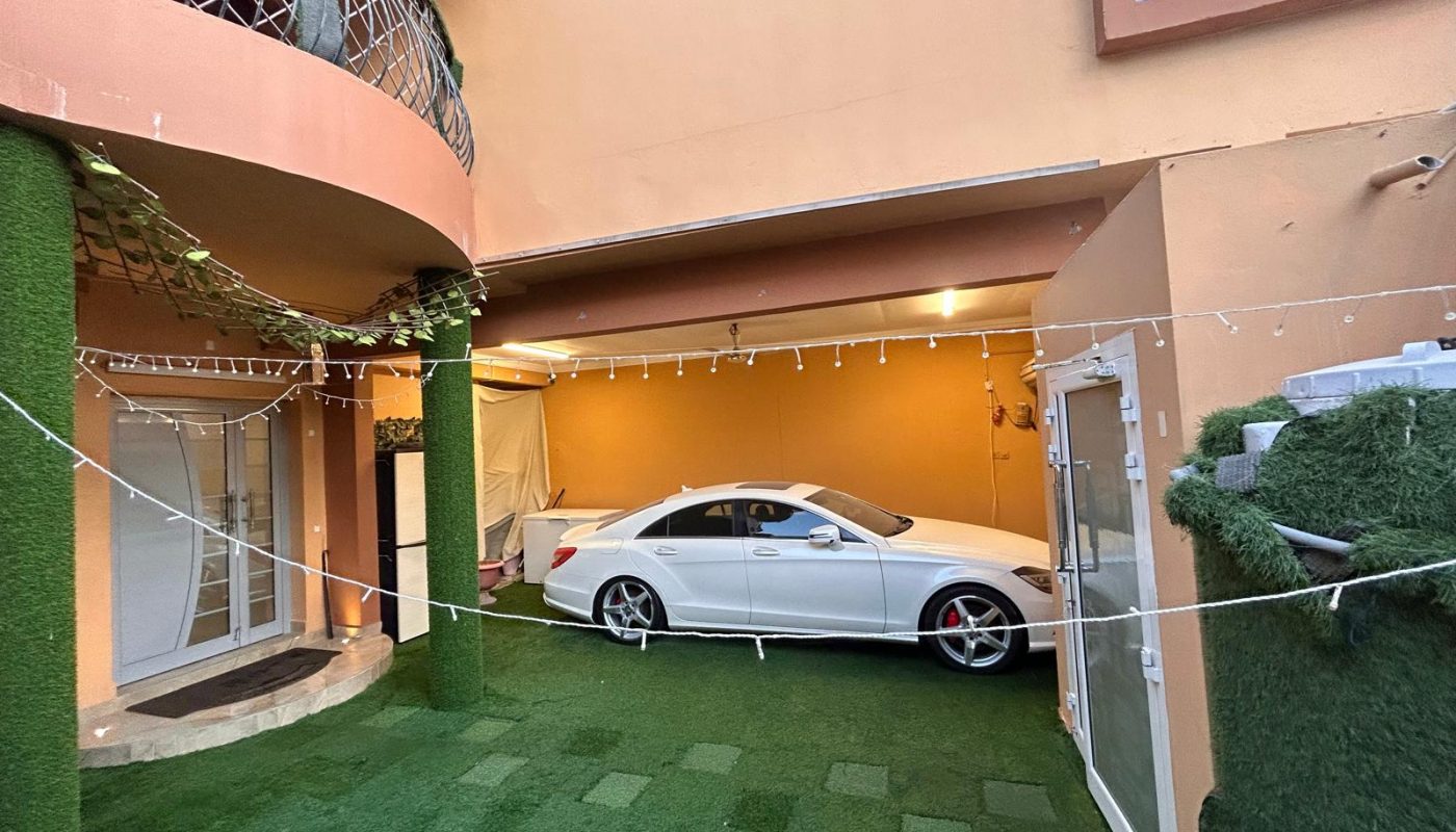 A white car is parked in the covered driveway of a peach-colored house with artificial grass covering the ground and pillars, and string lights hanging overhead.