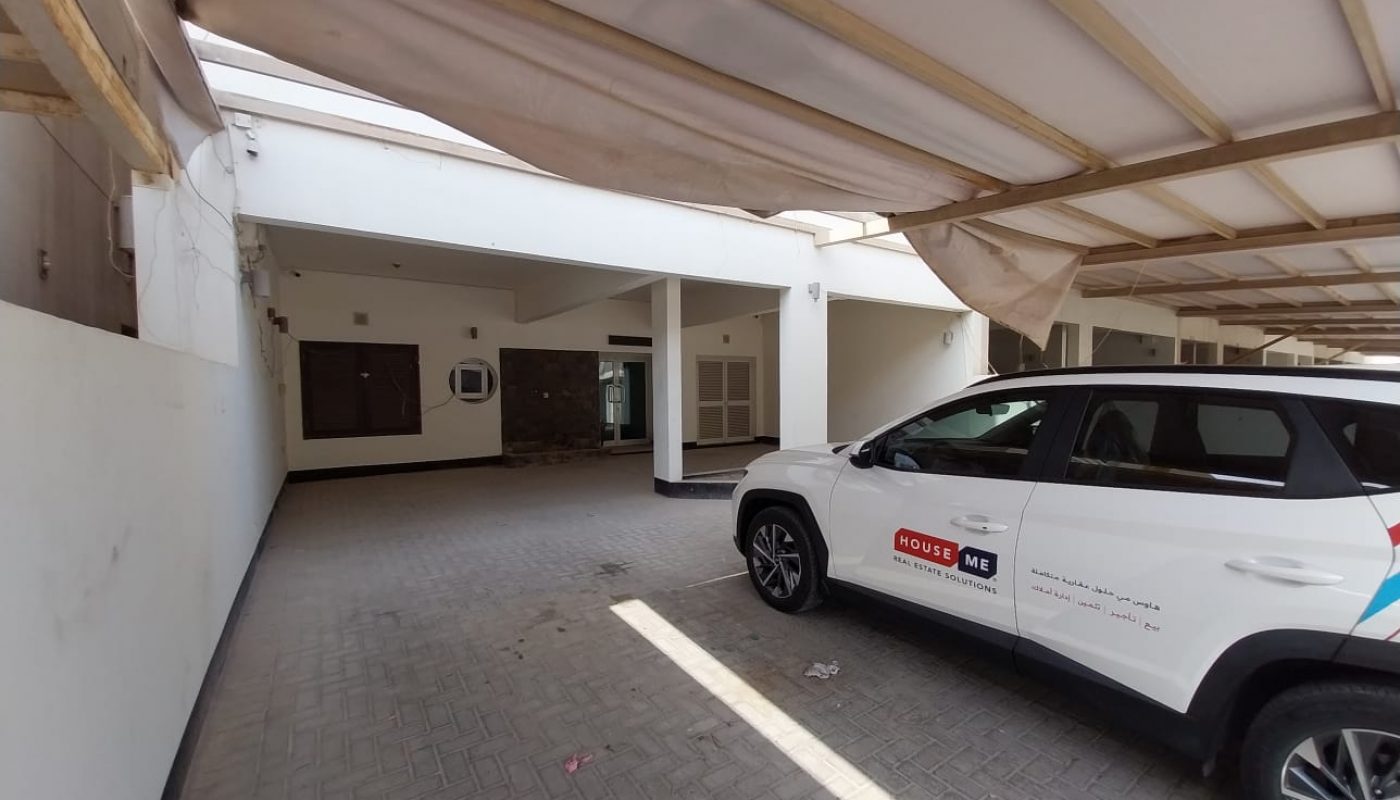 A white SUV with "House Me" branding is parked in a covered, tiled driveway next to a white building with several windows and doors.