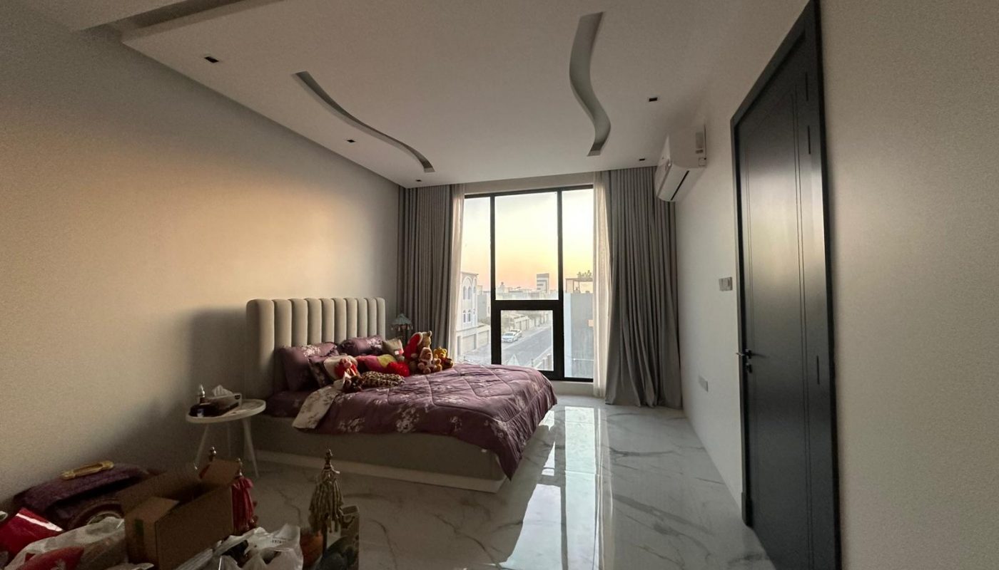 A modern bedroom with a large bed, purple bedding, floor-to-ceiling window, marble floor, and various items placed on the floor and side table.