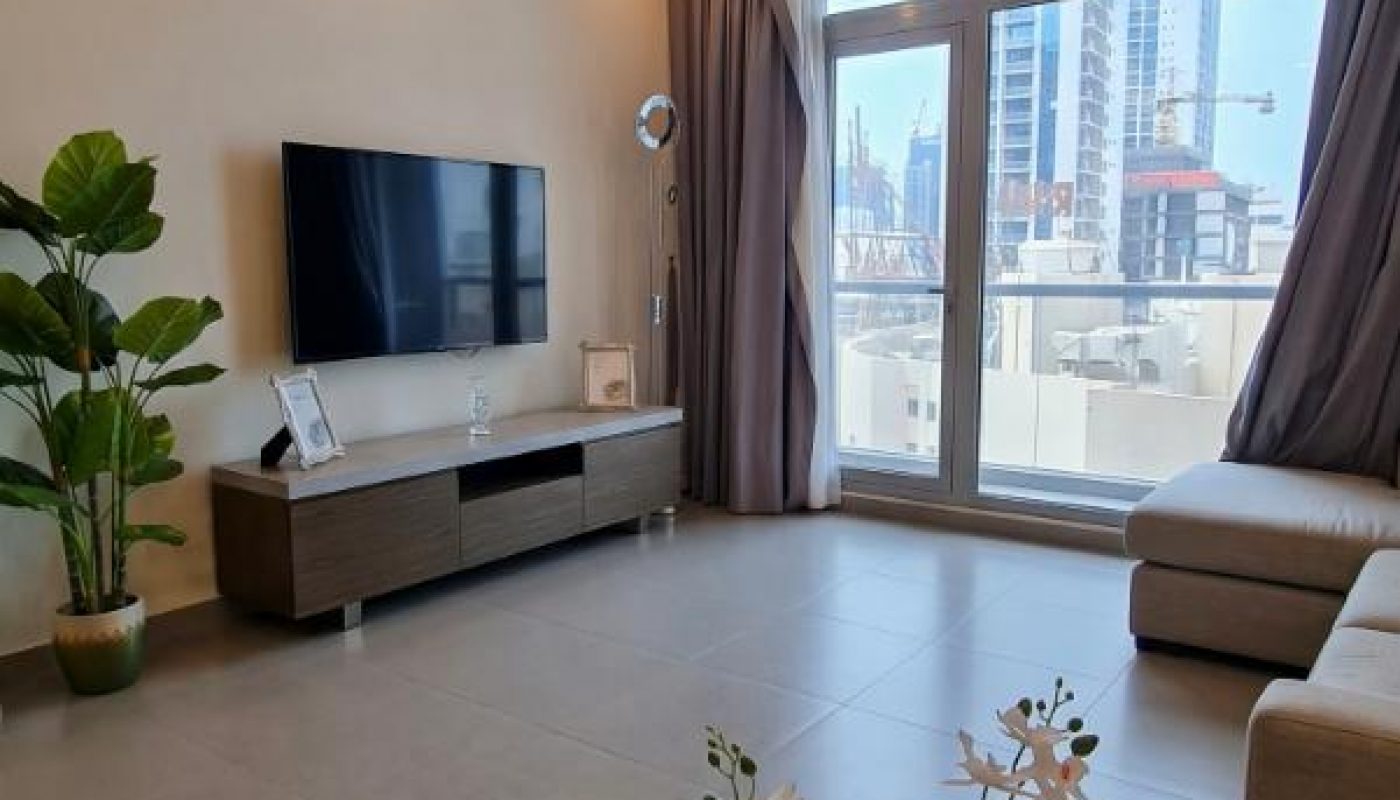 Modern living room with a wall-mounted TV, a low console, large windows showing city buildings, a potted plant, and a white orchid in the foreground.