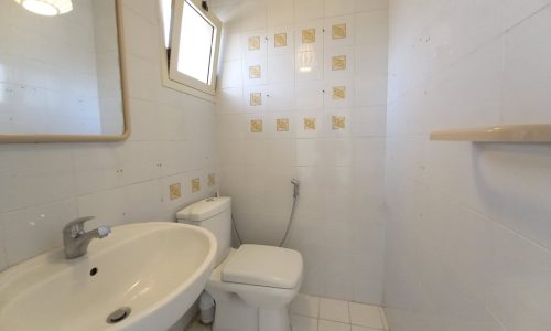 A small bathroom with white tiles, a sink with a faucet, a mirror, a toilet, and a bidet spray next to the toilet. A window near the ceiling provides natural light. A shelf is mounted on the wall.