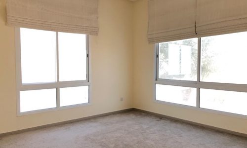 A room with light-colored walls and carpet, featuring two large windows with white blinds, allowing natural light to enter.