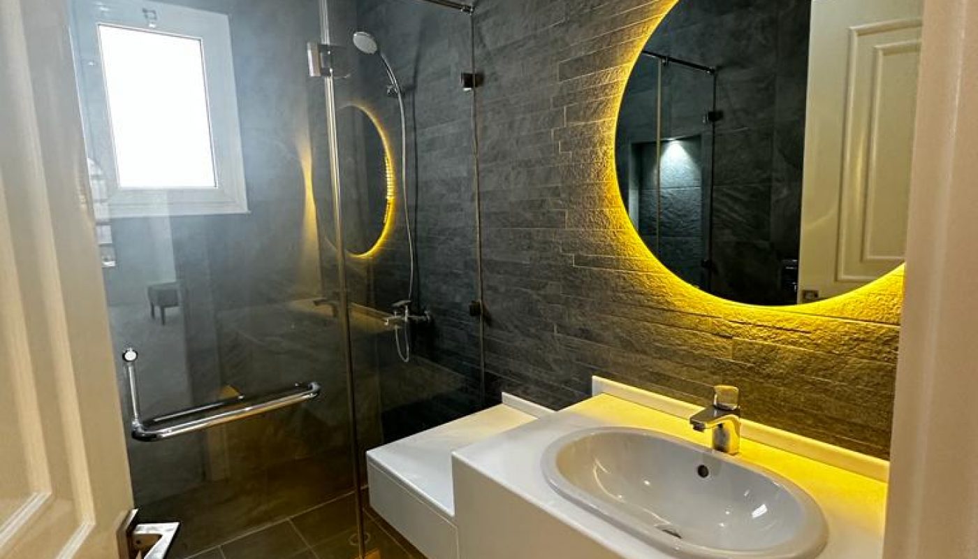 Modern bathroom with a walk-in glass shower, white sink, round mirror with yellow backlight, dark stone accent wall, and brown floor tiles.