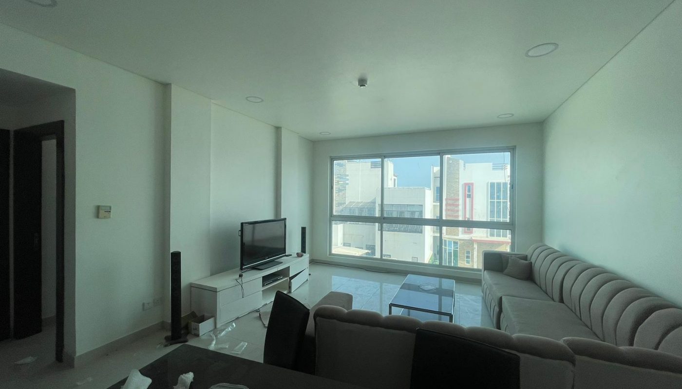 A modern living room with a large window, TV on a stand, sectional sofa, glass coffee table, and some clutter on the dining table in the foreground.