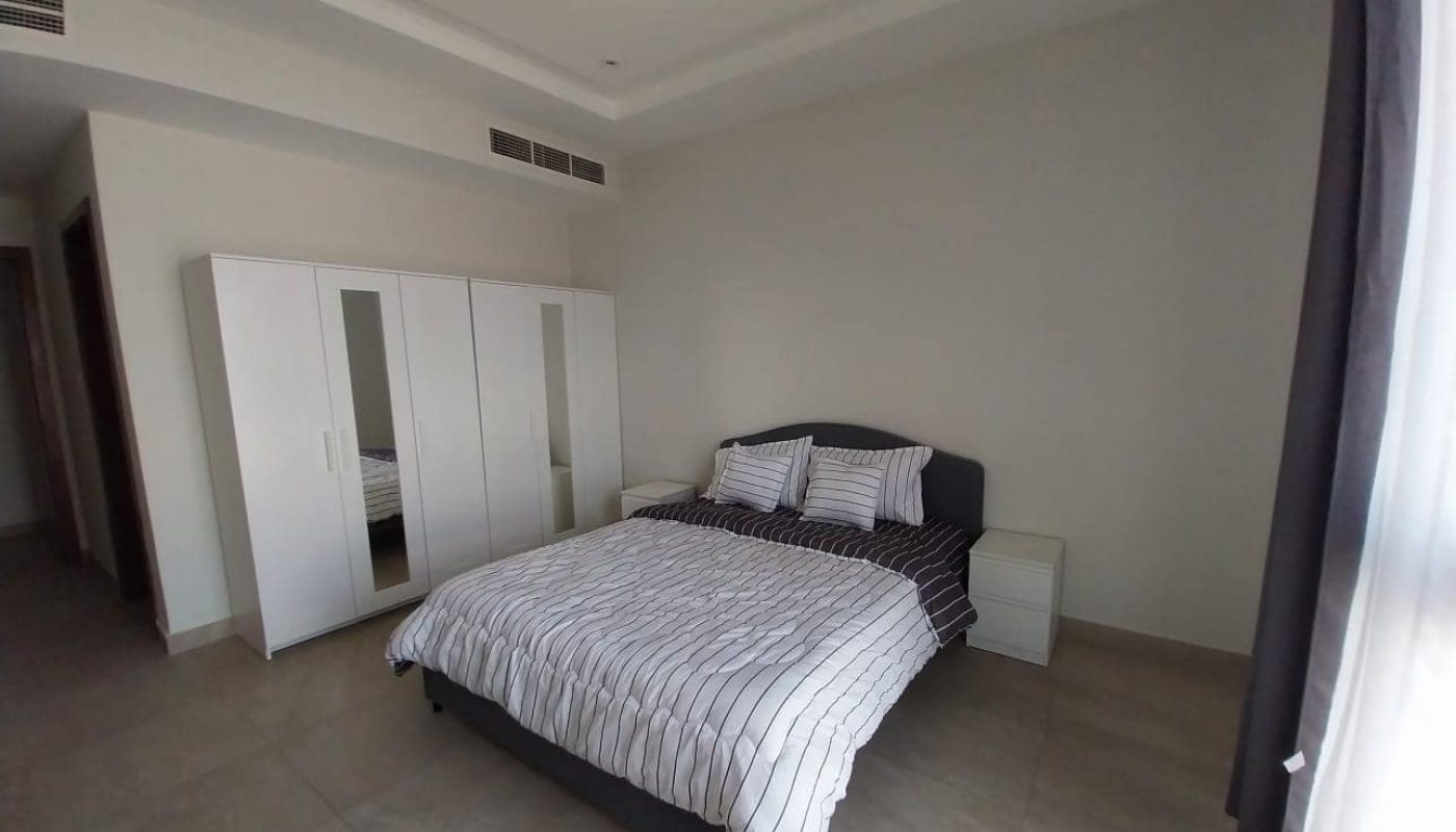 This flat for rent in Hidd features a cozy bedroom with a double bed, two bedside tables, and a wardrobe with mirrored doors. The room boasts beige tiled flooring and a large window adorned with curtains.