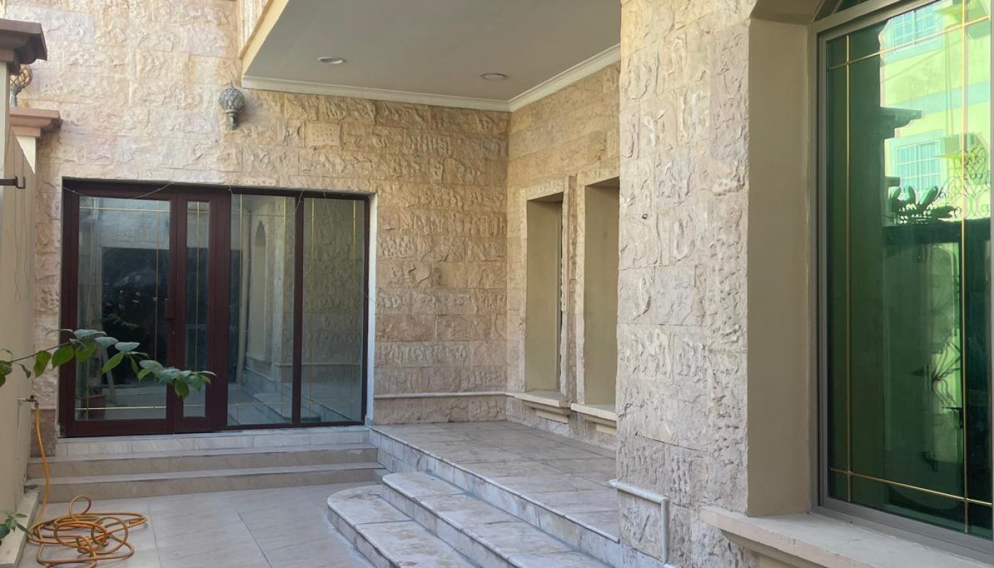A tiled outdoor patio with stone walls, steps leading to a doorway, large windows, and potted plants along the sides.