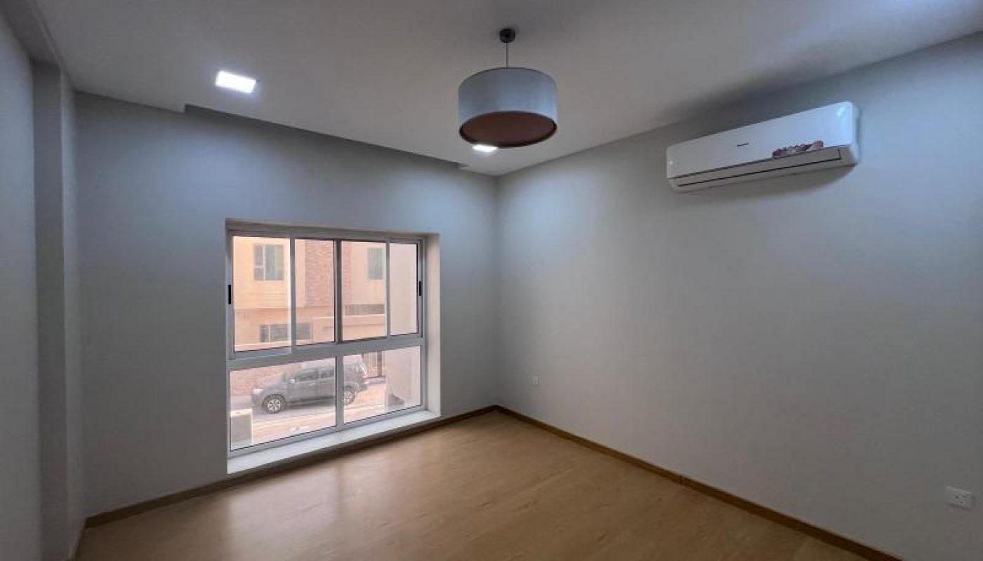 Empty room with light-colored walls, wooden floor, large window, ceiling light fixture, and wall-mounted air conditioner.