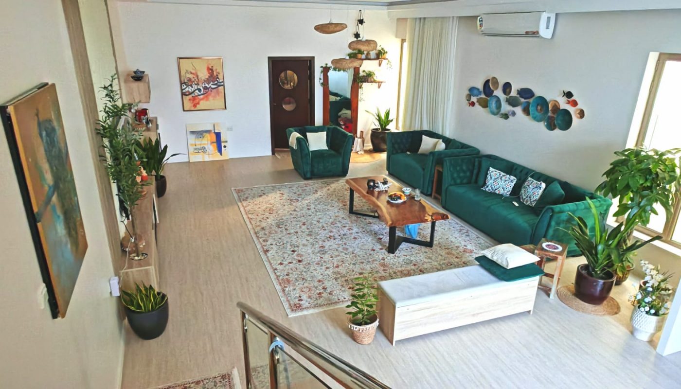 Modern living room with green sofas, a patterned rug, wooden coffee tables, wall art, numerous houseplants, and large windows letting in natural light.