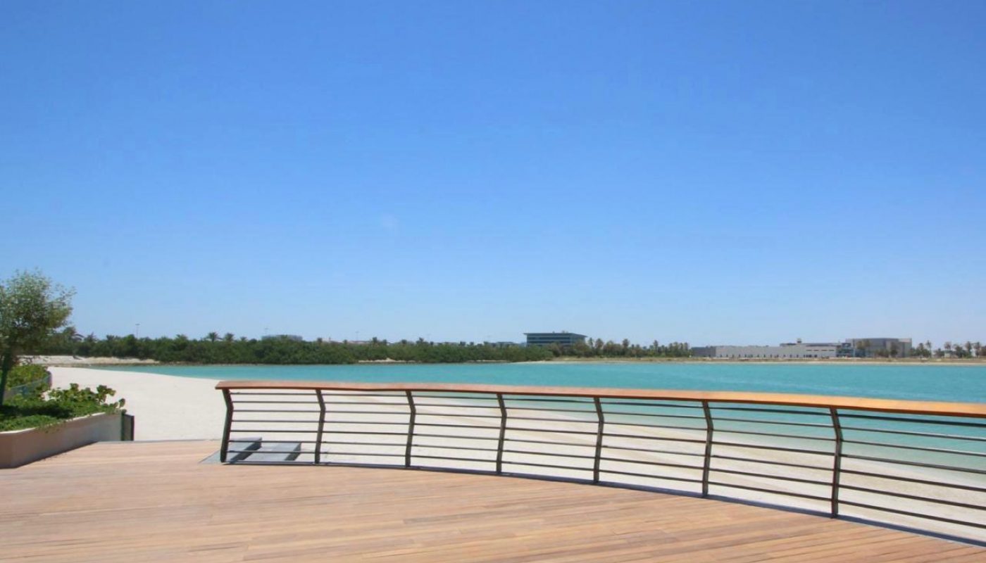 Wooden deck with metal railing overlooks a calm body of water, sandy shore, and distant buildings under a clear blue sky.