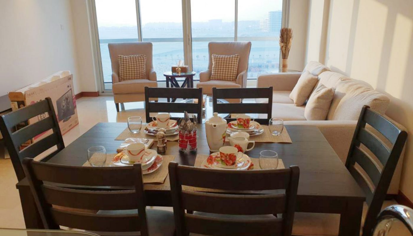 A dining table set for four with plates of food and drinks, in a bright living room with large windows, two armchairs, and a sofa.
