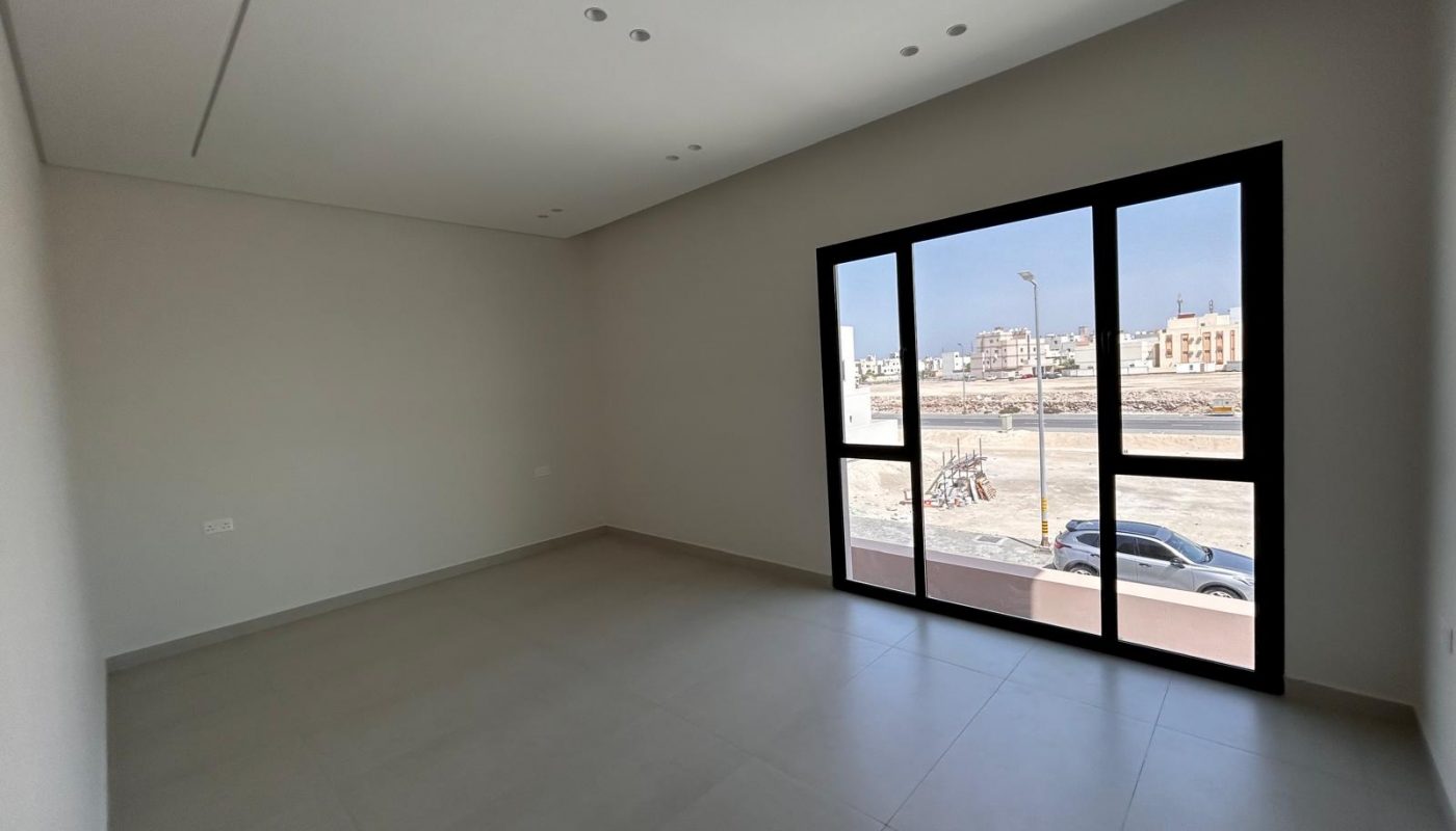 Empty, unfurnished room with light-colored tiled floor, plain walls, and large black-framed window showing urban buildings and parked cars outside.