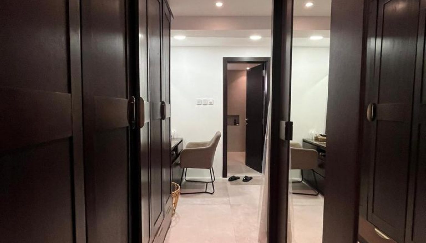 A modern walk-in closet with dark wooden wardrobes, a large mirror, a chair at a desk, and light-colored tiled flooring under ceiling lights.