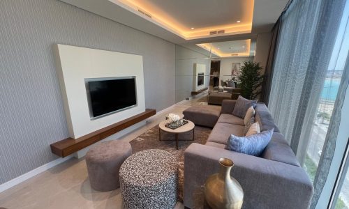 Modern living room in a 2 bedroom apartment with a large sectional couch, rounded ottomans, a wall-mounted TV, and expansive windows with city views.